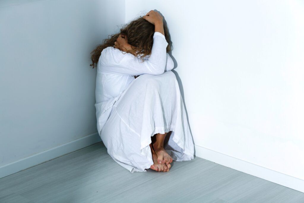 a woman with schizophrenia hiding in a corner of a room as a result of hearing voices, showing the need to treat schizophrenia with the proper approach